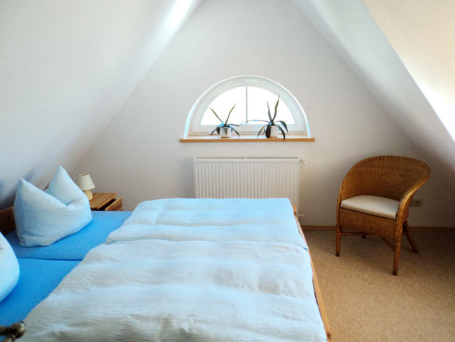 Schlafzimmer der Oberen Ferienwohnung mit Doppelbett, Nachtschrank, Stuhl und rundem Gaubenfenster